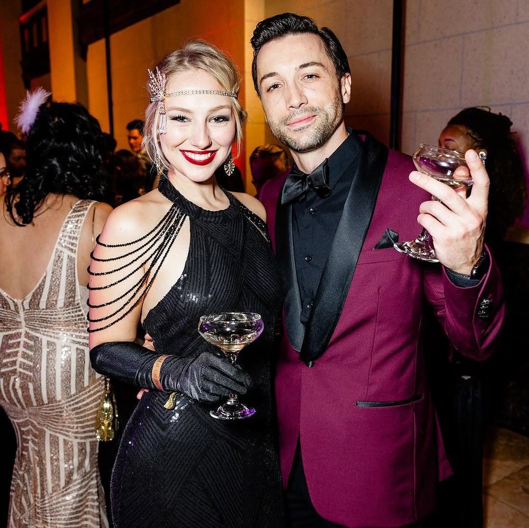 Man in maroon tuxedo and woman in black flapper dress holding cocktail glasses.