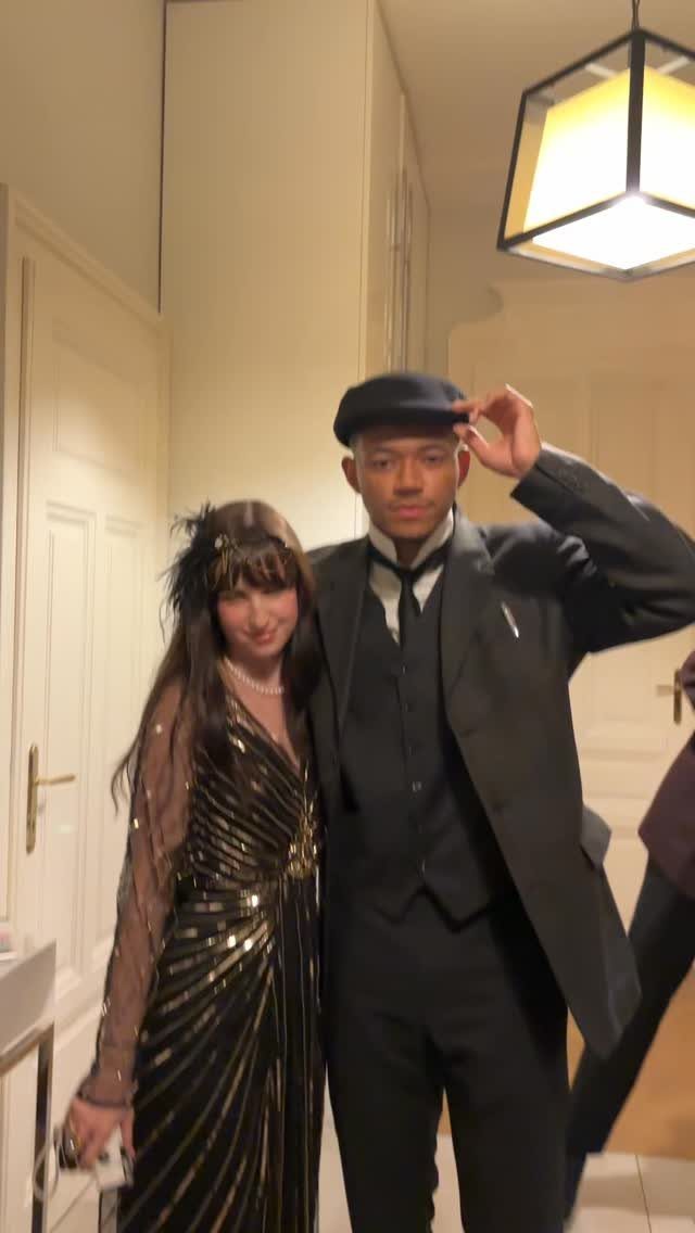 Man in a newsboy cap and suit beside woman in sequined dress and headpiece.