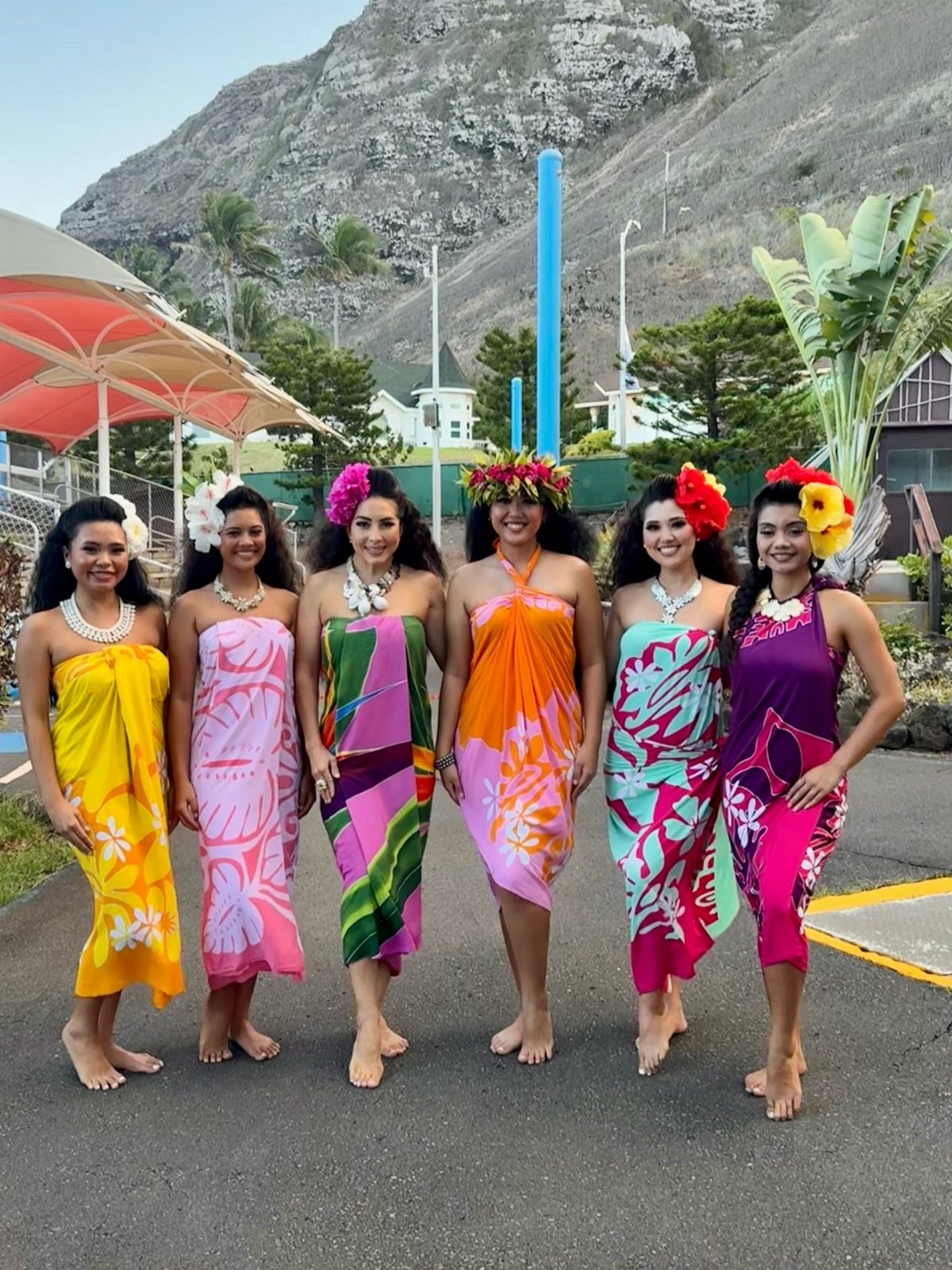 Six women wearing colorful tropical sarongs and floral headpieces standing outdoors near mountains.