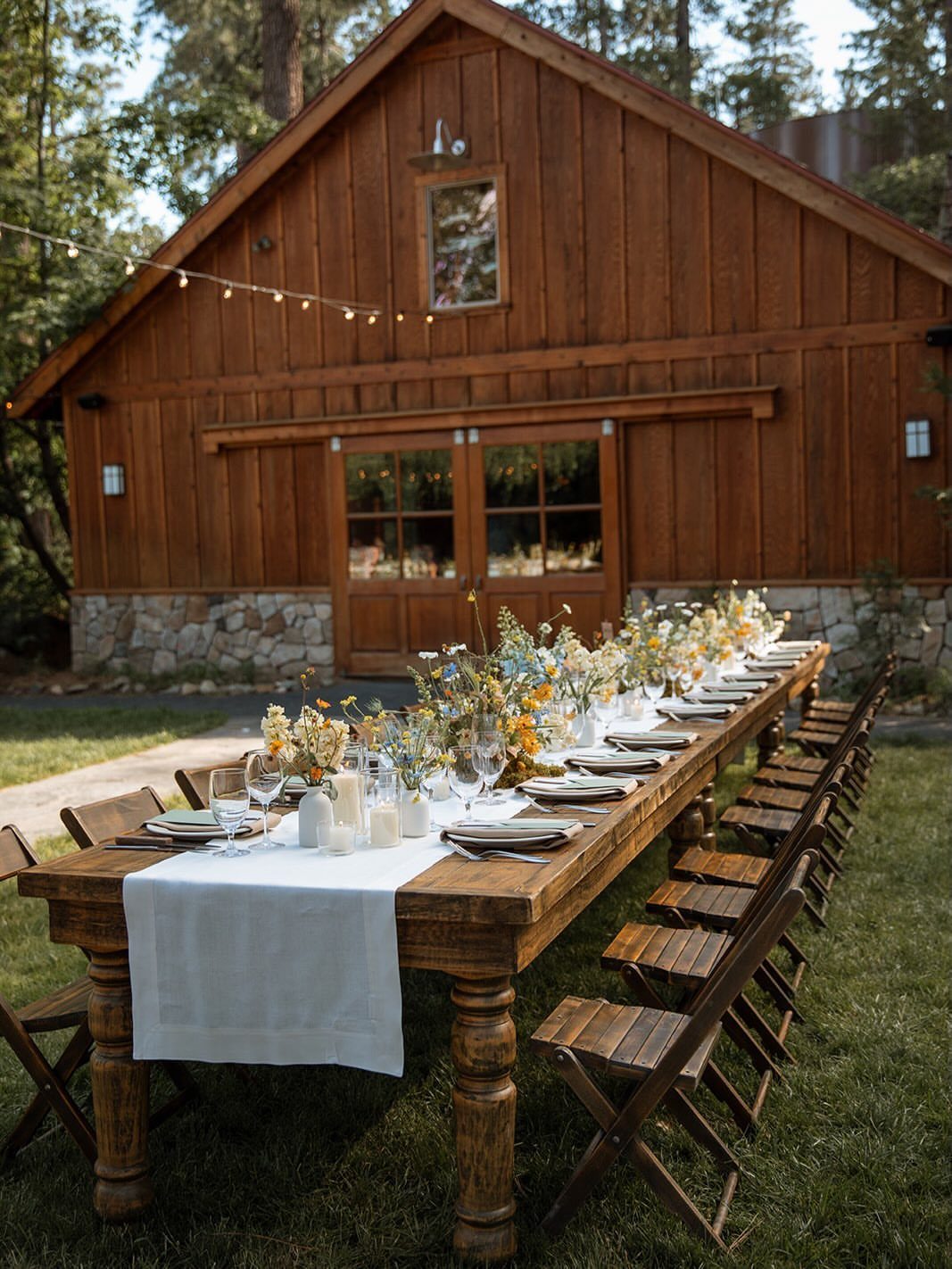 Long wooden dining table with wildflower centerpieces set outdoors before a rustic barn.