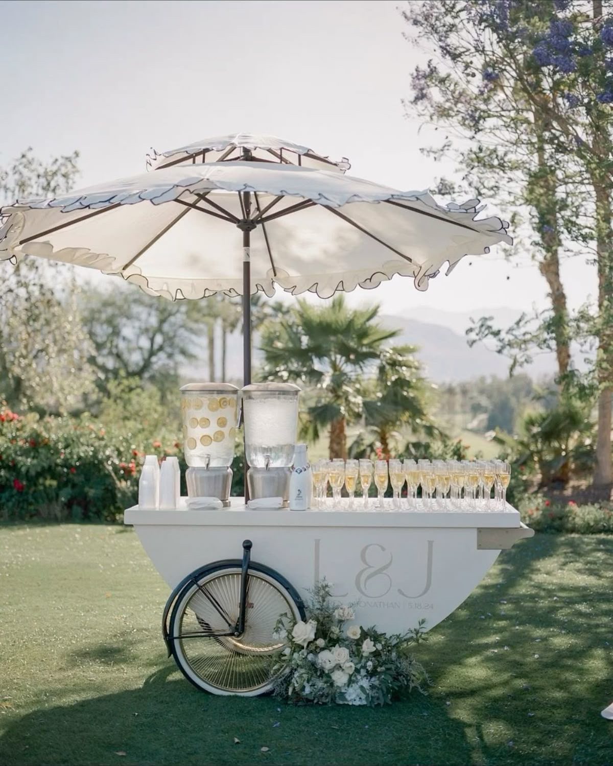 White beverage cart with champagne flutes and a white umbrella on a lawn