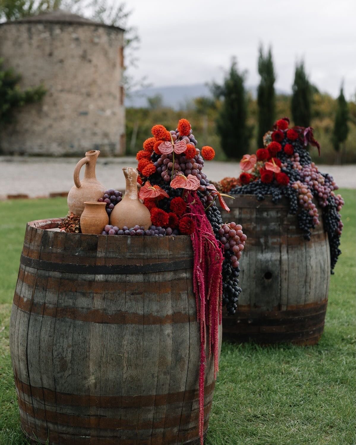 Wooden wine barrels decorated with autumn flowers, grapes, and terracotta jugs on grass.