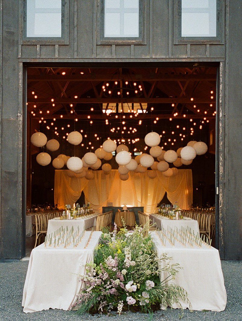 White paper lanterns and string lights hanging over a decorated wedding reception table.