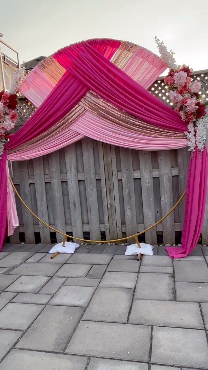 Gold ceremony arch draped in pink fabric and flowers against a wooden fence.