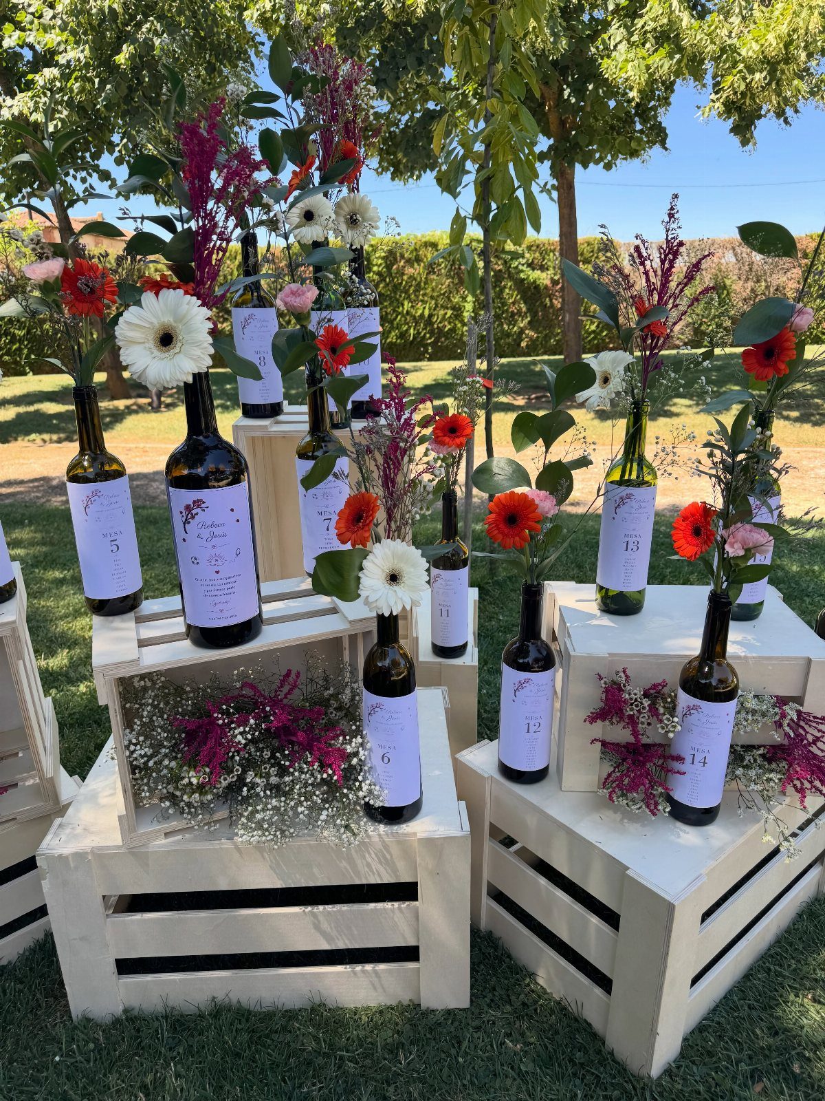 Wine bottles used as flower vases arranged on white wooden crates outdoors.