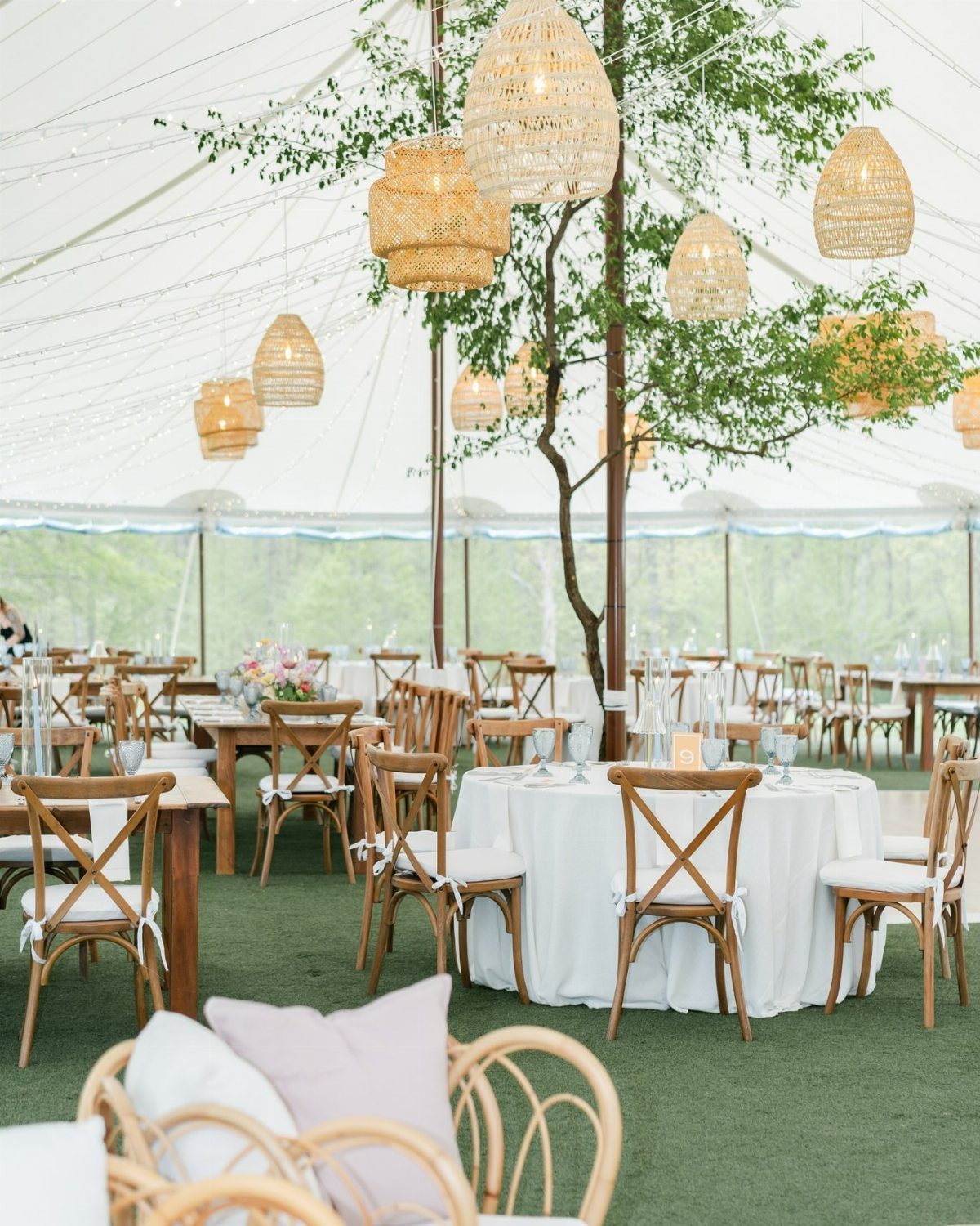 Wedding reception under a white tent with woven wicker pendant lamps and wooden furniture.