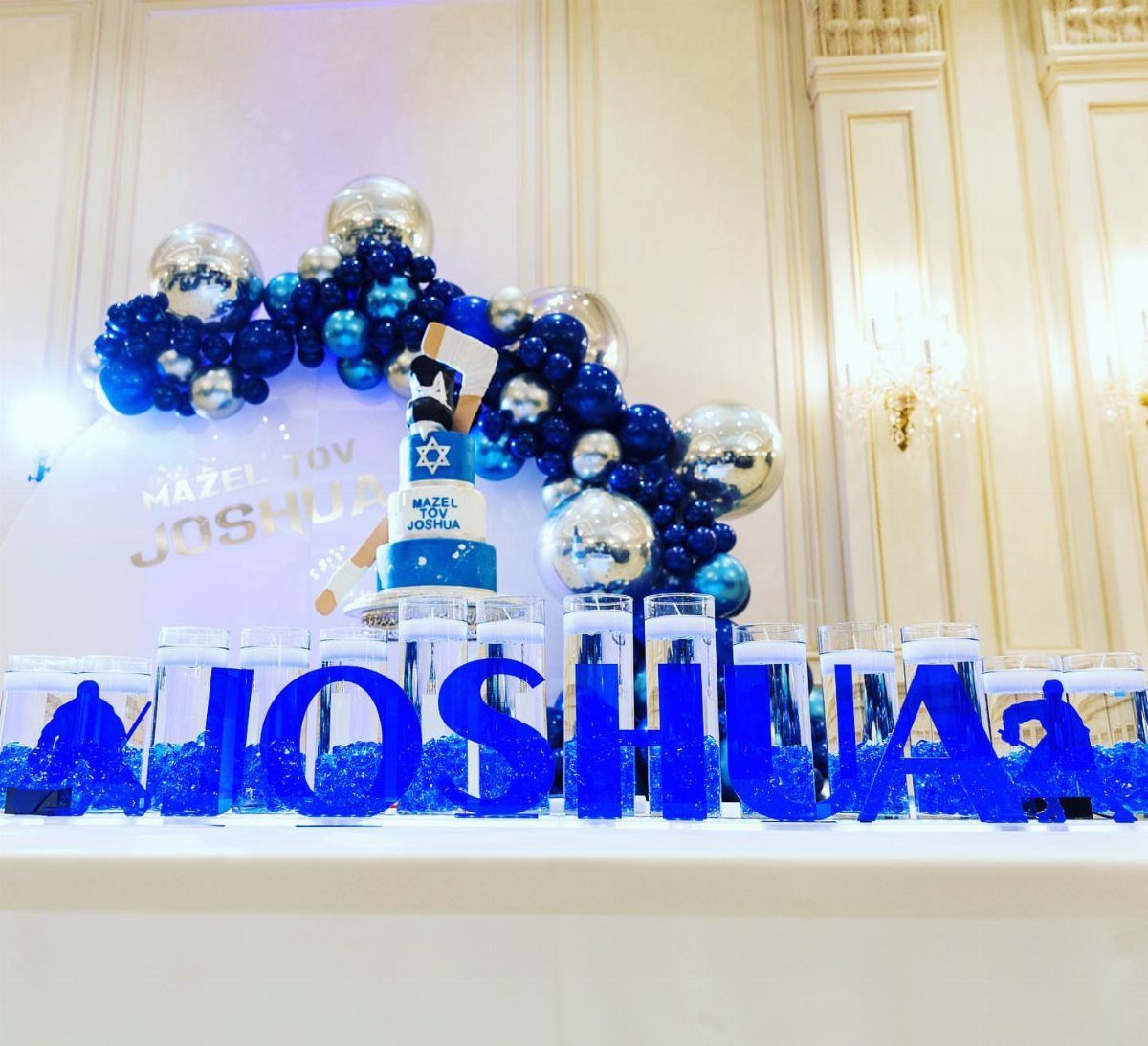 Bar Mitzvah dessert table with blue balloons, a themed cake, and blue letter cylinders.