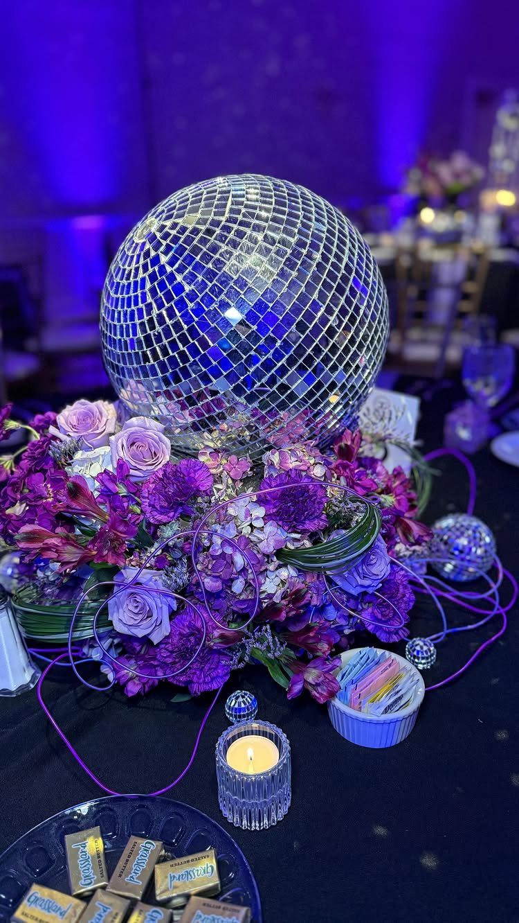Disco ball centerpiece resting on purple floral arrangements with a small candle nearby.