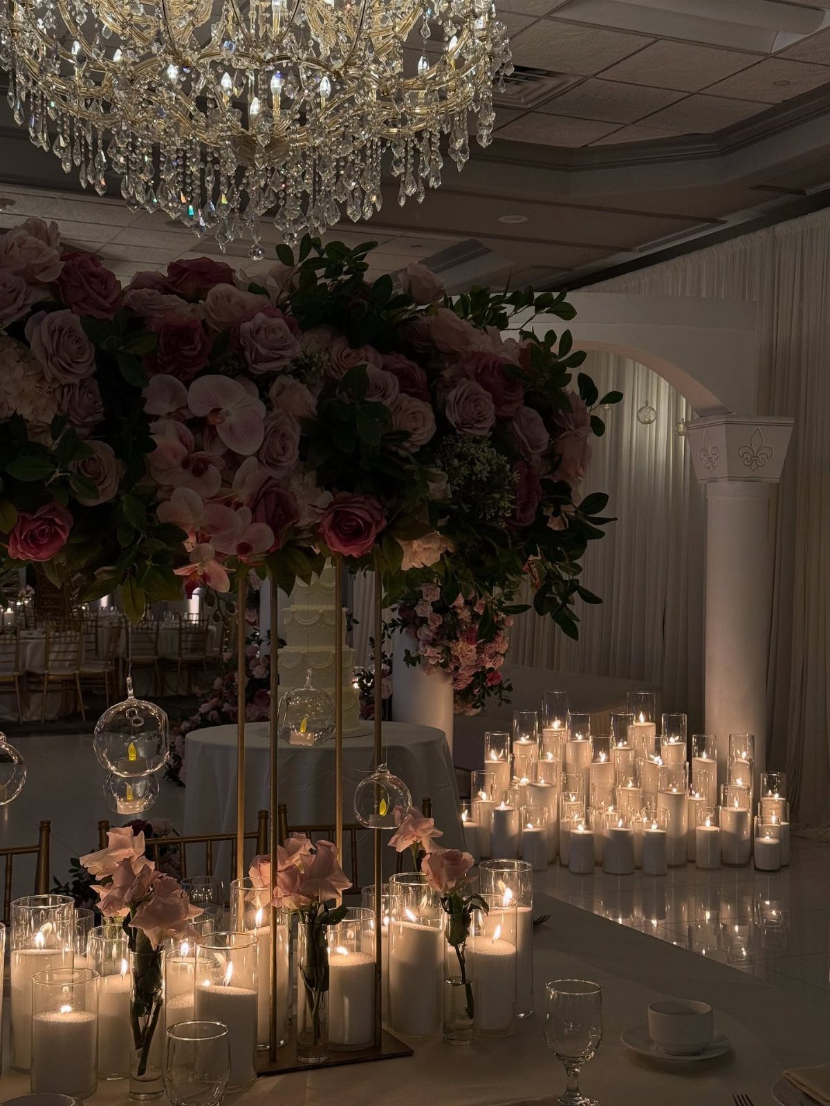 Pink rose floral arrangement and many white pillar candles in glass cylinders.