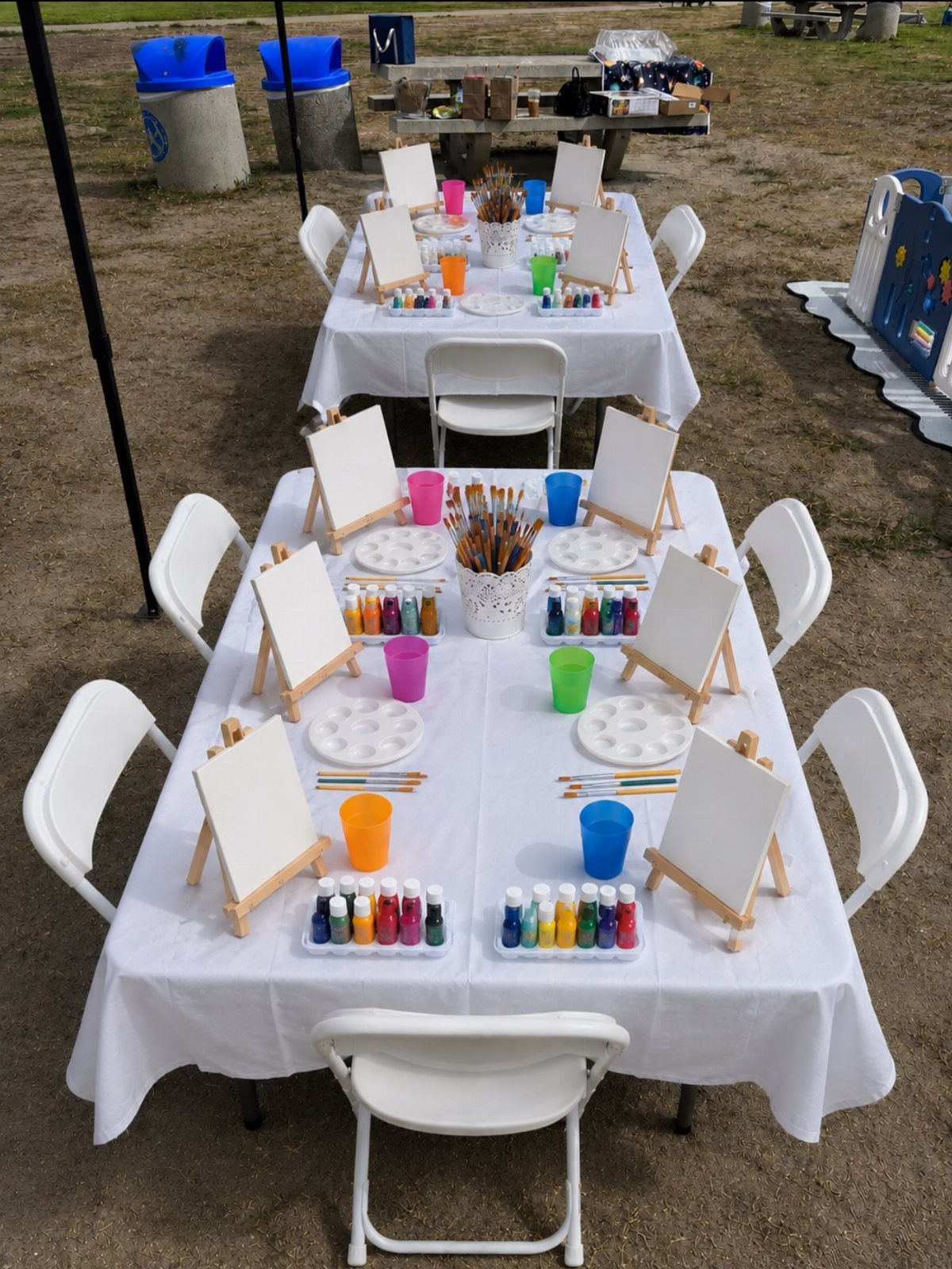 Art stations with small wooden easels, blank canvases, and colorful paints on white tables.