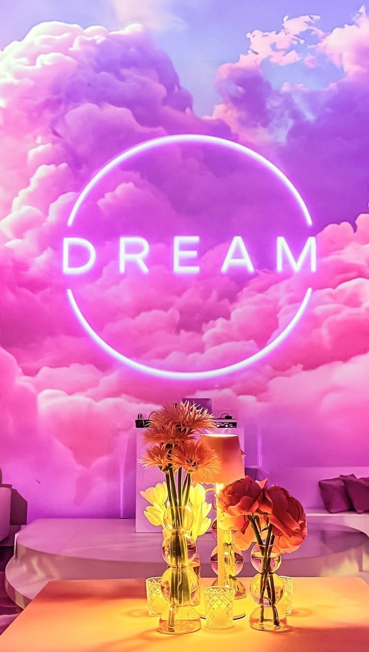 Neon DREAM sign in a circle against a purple cloud backdrop with flowers.