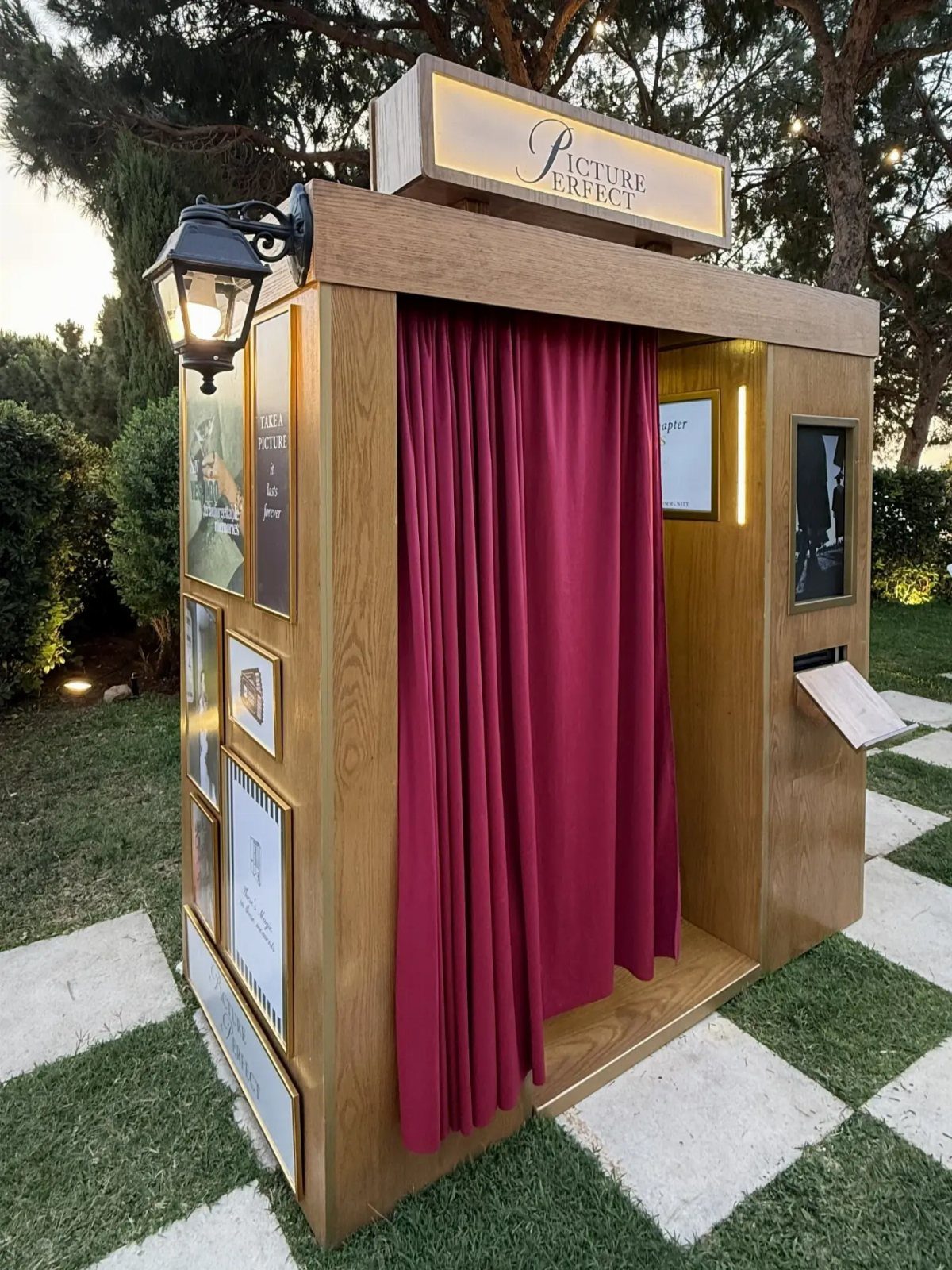 Wooden outdoor photo booth with red velvet curtains and illuminated sign on grass.