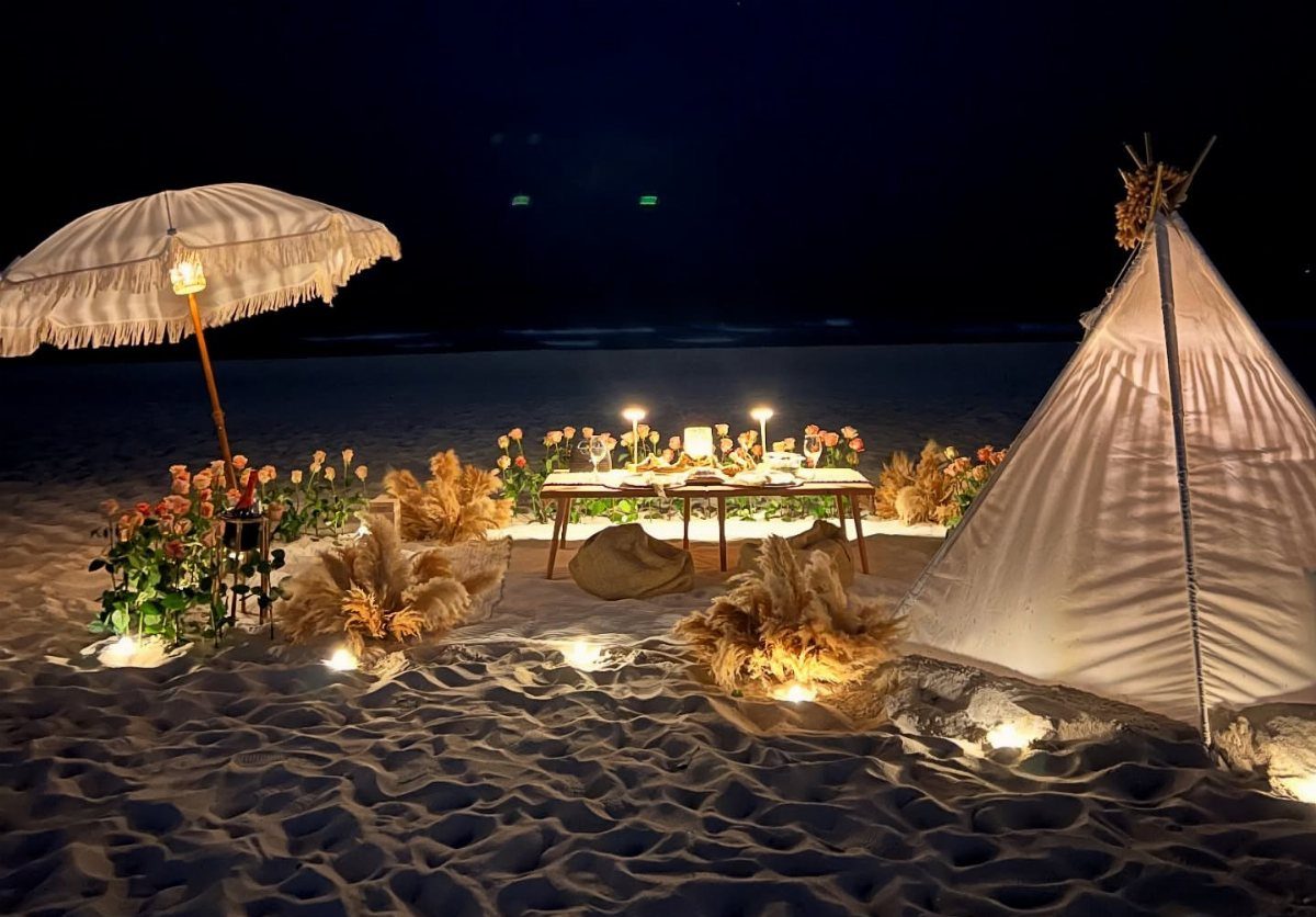 Romantic beach dinner setup with a teepee, umbrella, and candles on white sand