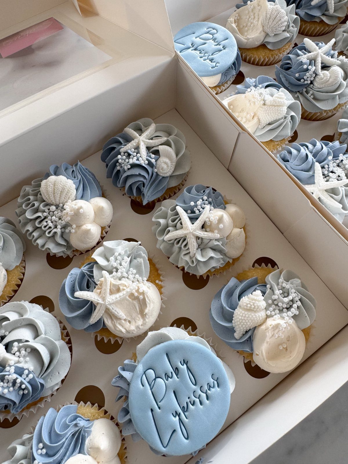 Beach-themed cupcakes with blue and white frosting, edible seashells, and starfish in a box.