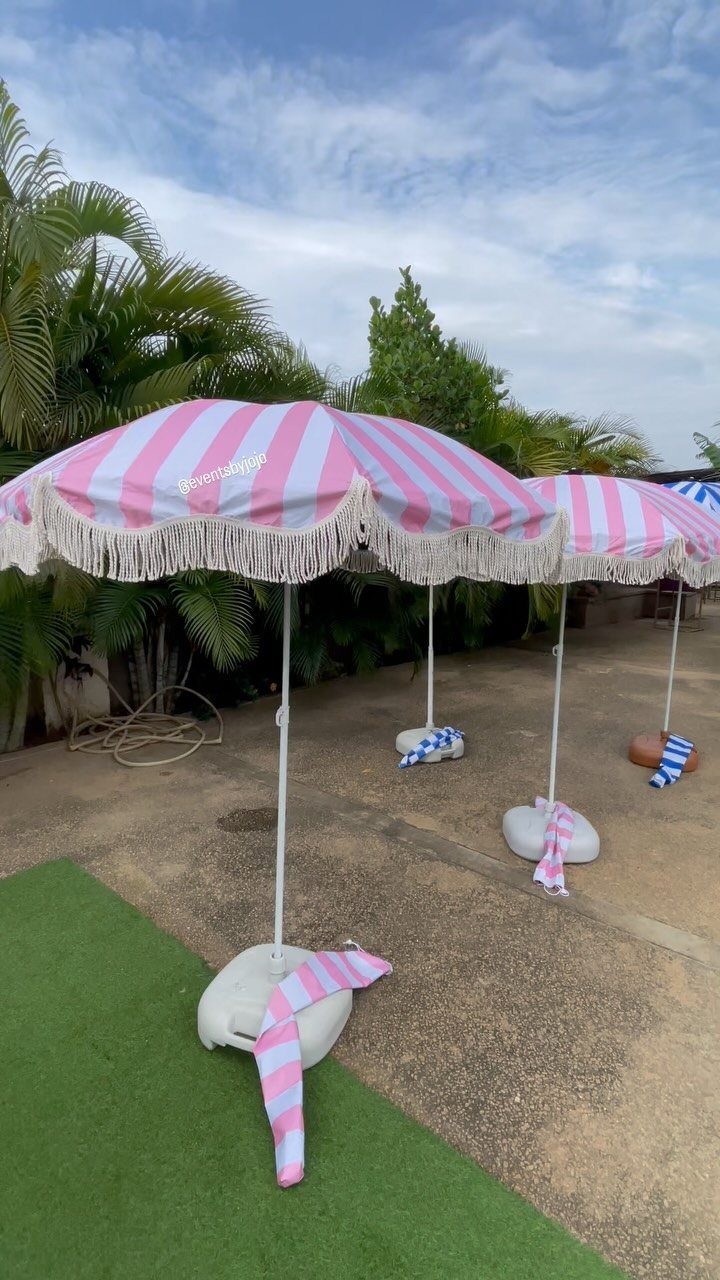 Pink and white striped beach umbrellas with fringe standing on sand and turf.
