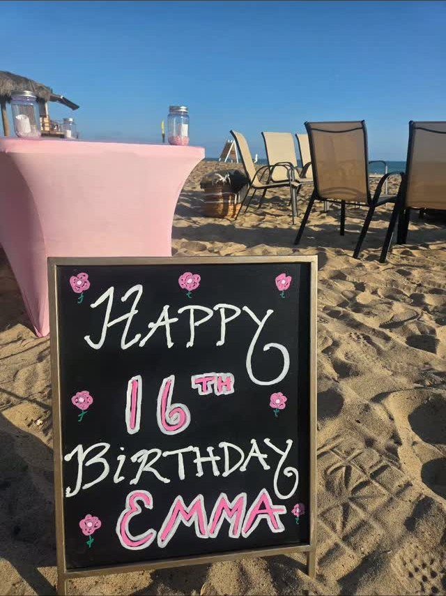 Birthday chalkboard sign on a sandy beach with pink table and lounge chairs.