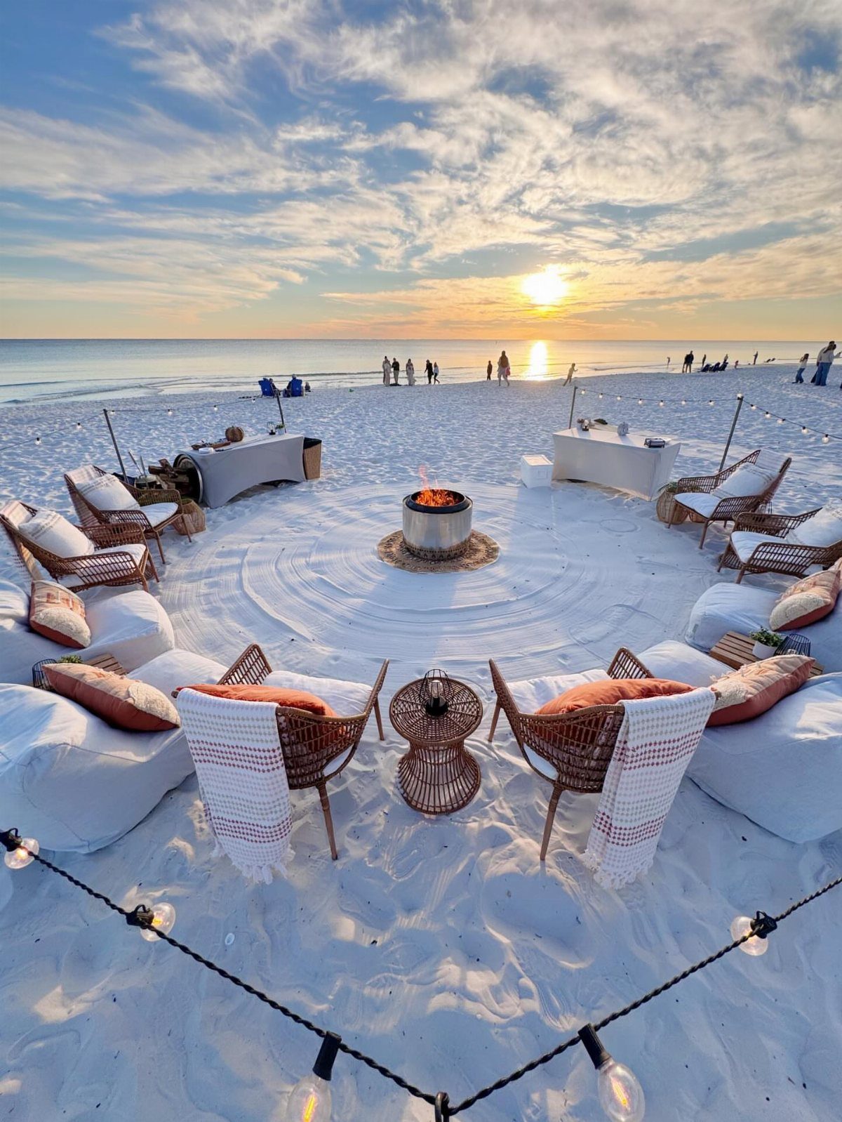Beach bonfire setup with wicker chairs and bean bags around a fire pit at sunset.