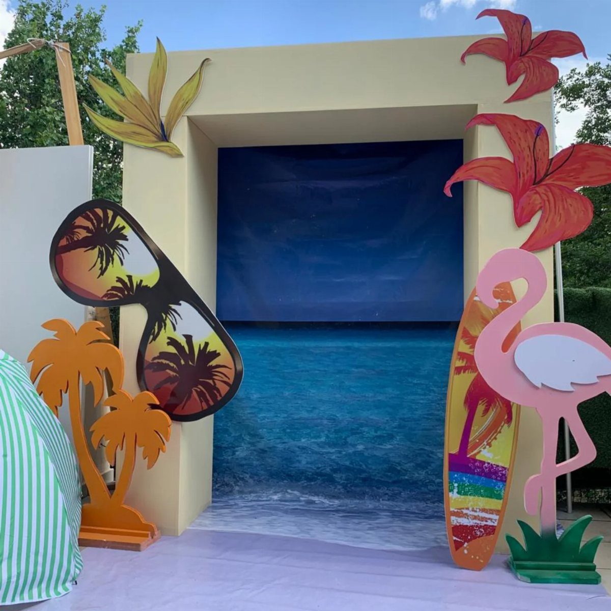 Beach-themed photo booth with ocean backdrop, flamingo, surfboard, and oversized sunglasses cutouts.