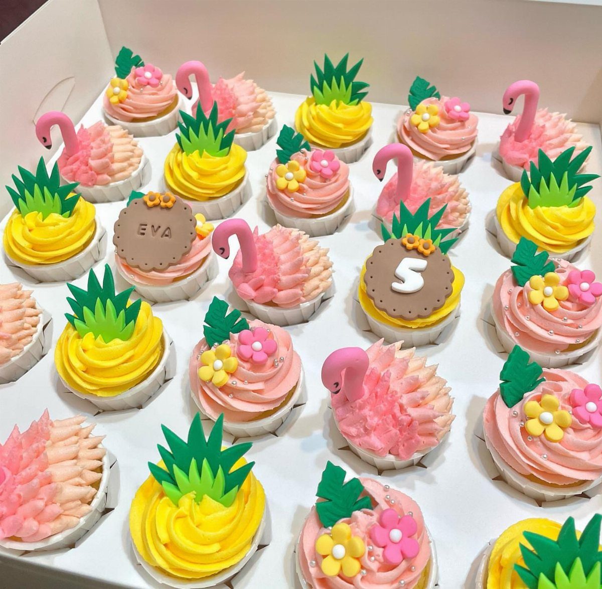 Assorted cupcakes with pink flamingo and yellow pineapple frosting in a white box.