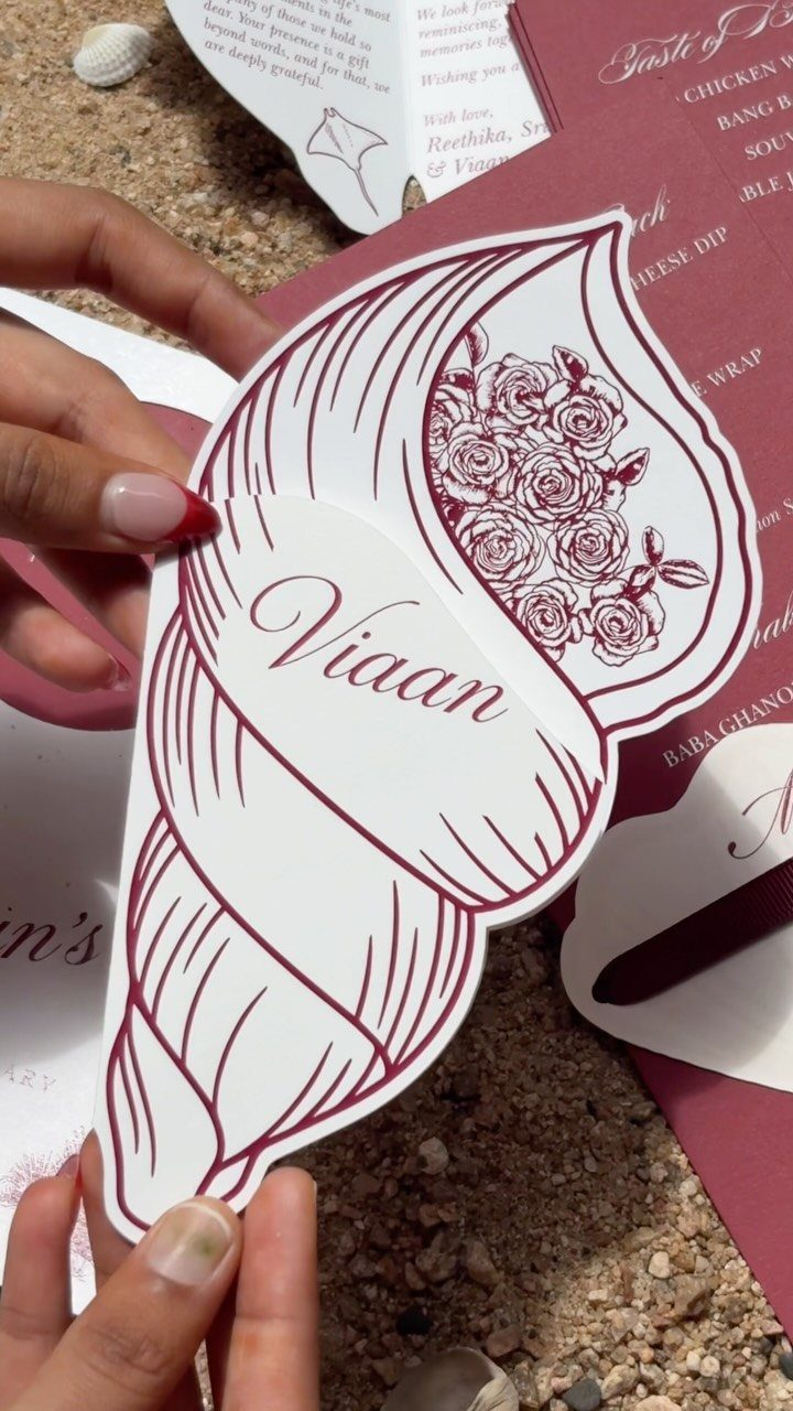 Seashell shaped place card with red floral art held over sandy beach background