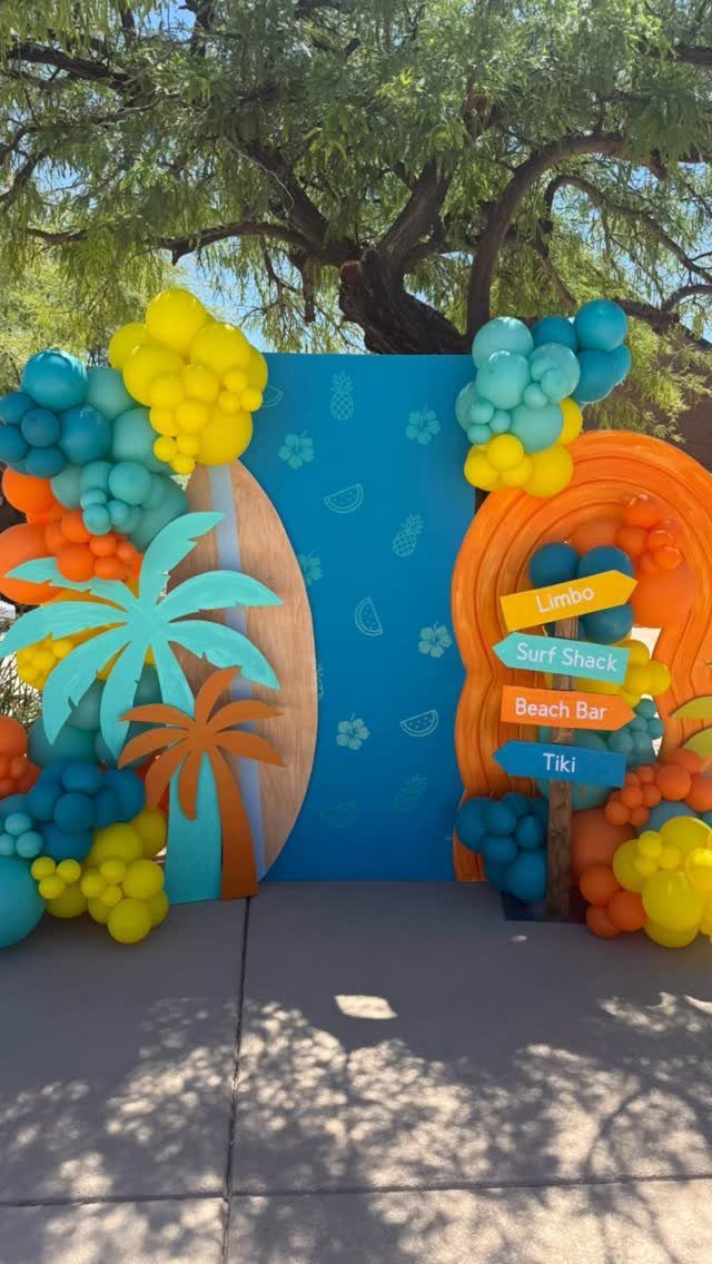 Beach party decoration with colorful balloon arches, palm tree cutout, and directional signpost.