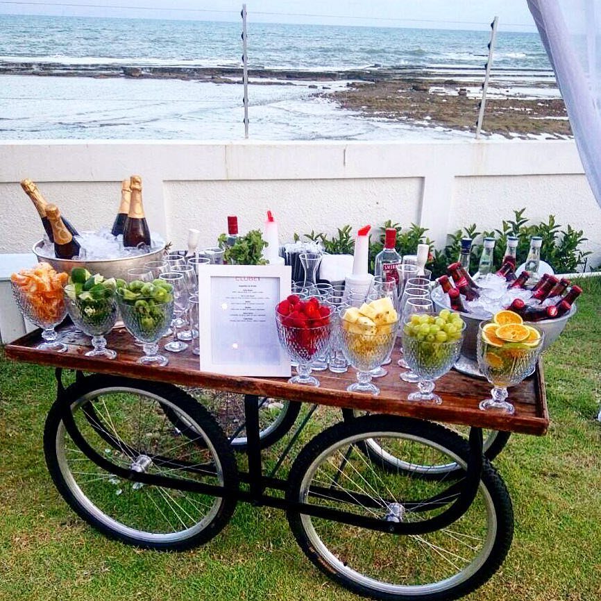 Wooden drink cart with champagne, bottled beverages, and fresh fruit bowls by the ocean.