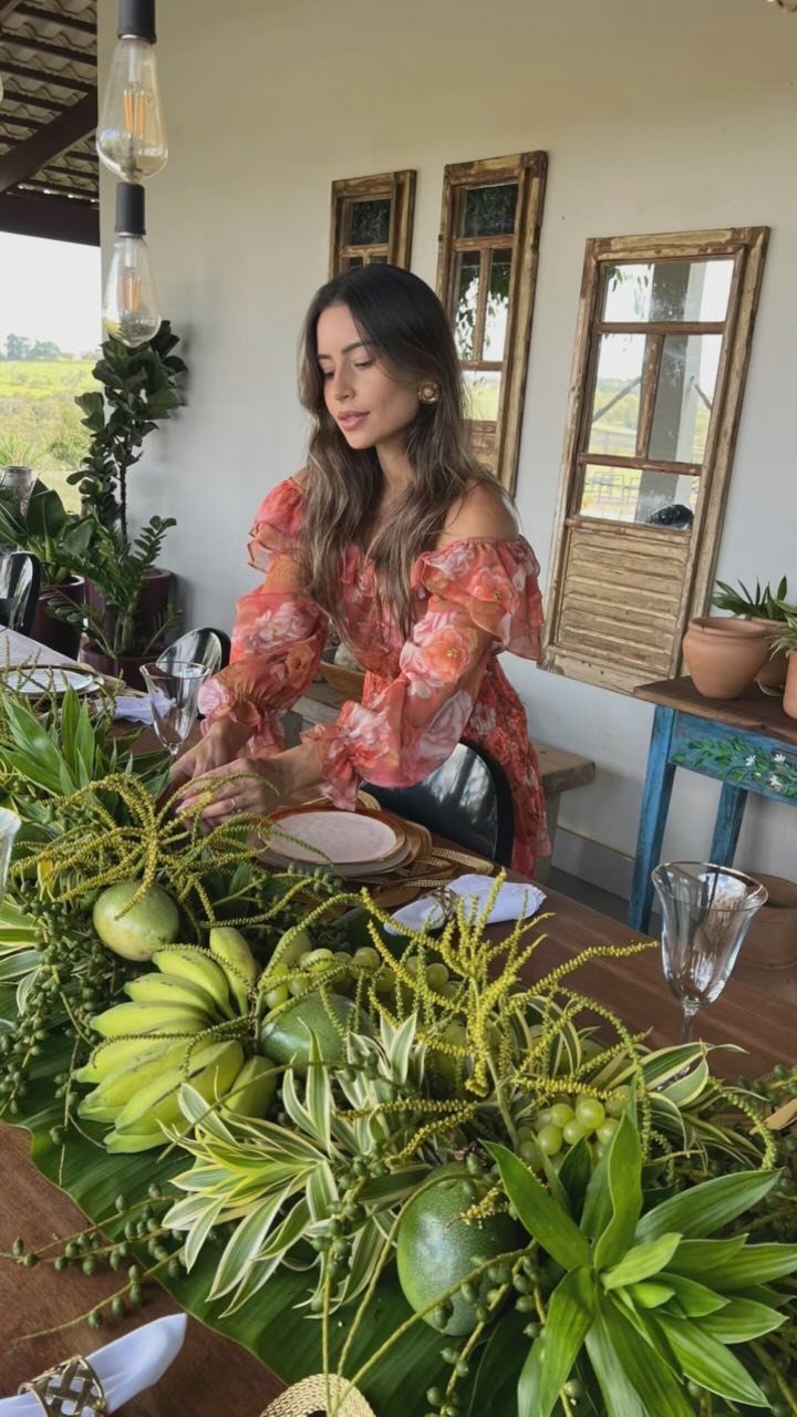 Woman arranging a tropical tablescape with bananas, coconuts, and green foliage on wood.
