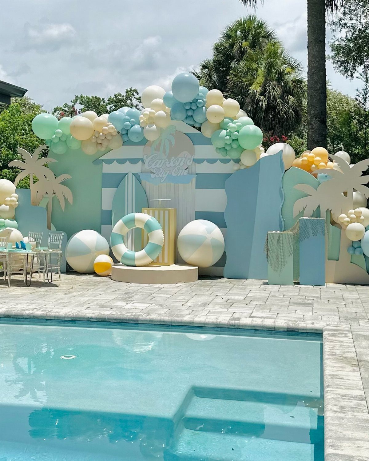 Beach themed party decorations with blue and cream balloons by a swimming pool.