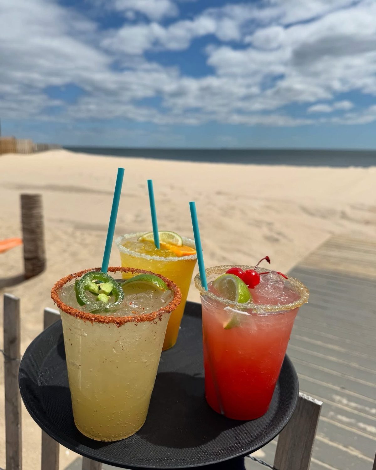 Three colorful beach cocktails with garnishes on a tray at a sandy beach.