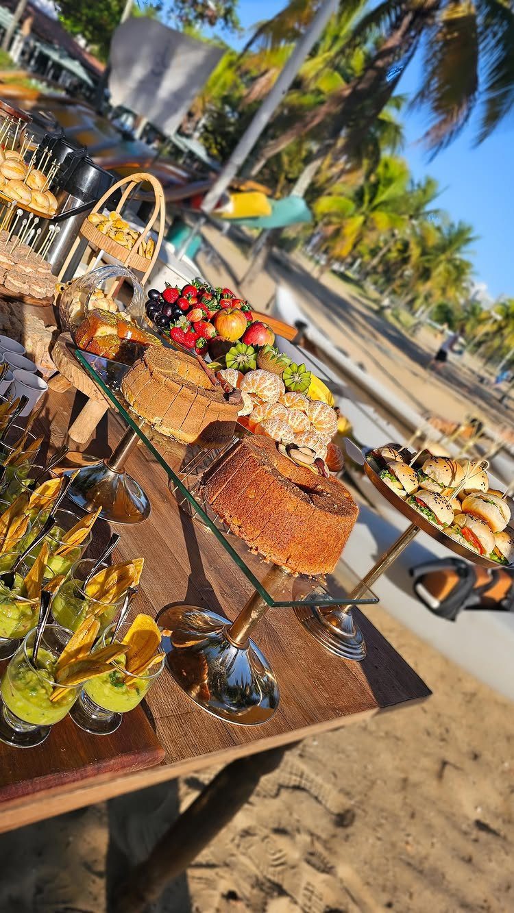 Buffet table on a beach with cakes, fruit, and appetizers on glass stands.