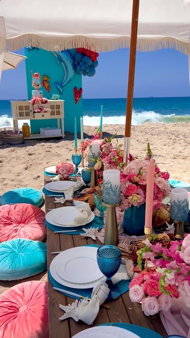 Beach party table setting with pink flowers, blue plates, and cushions on sand.