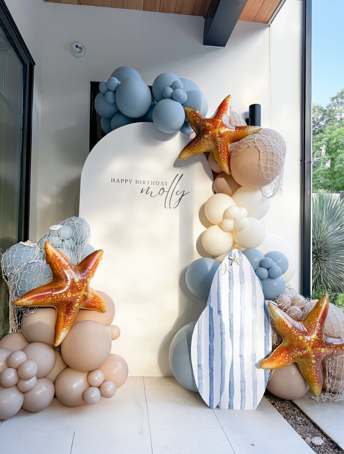 Beach themed birthday backdrop with blue balloons, orange starfish, and a striped surfboard.