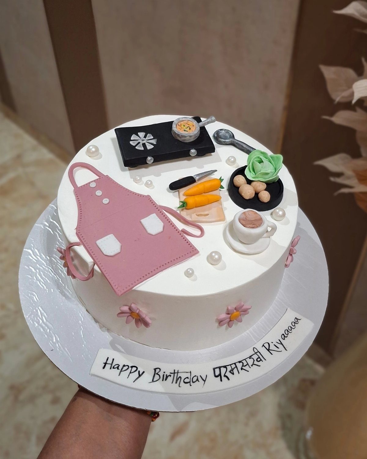 White birthday cake decorated with edible miniatures of a pink apron and cooking utensils.