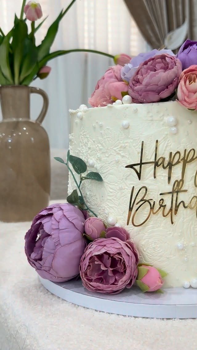 White birthday cake decorated with purple and pink peonies and gold lettering.