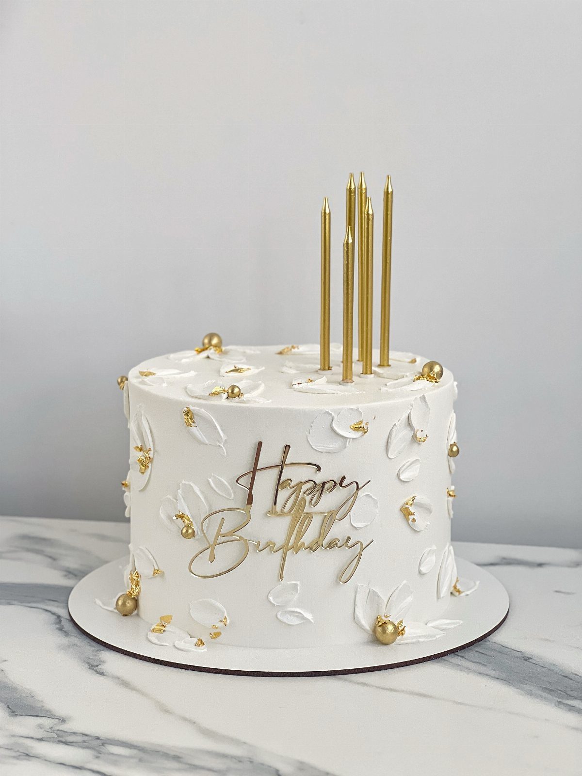 White frosted birthday cake with gold candles, gold pearls, and white petal decorations.