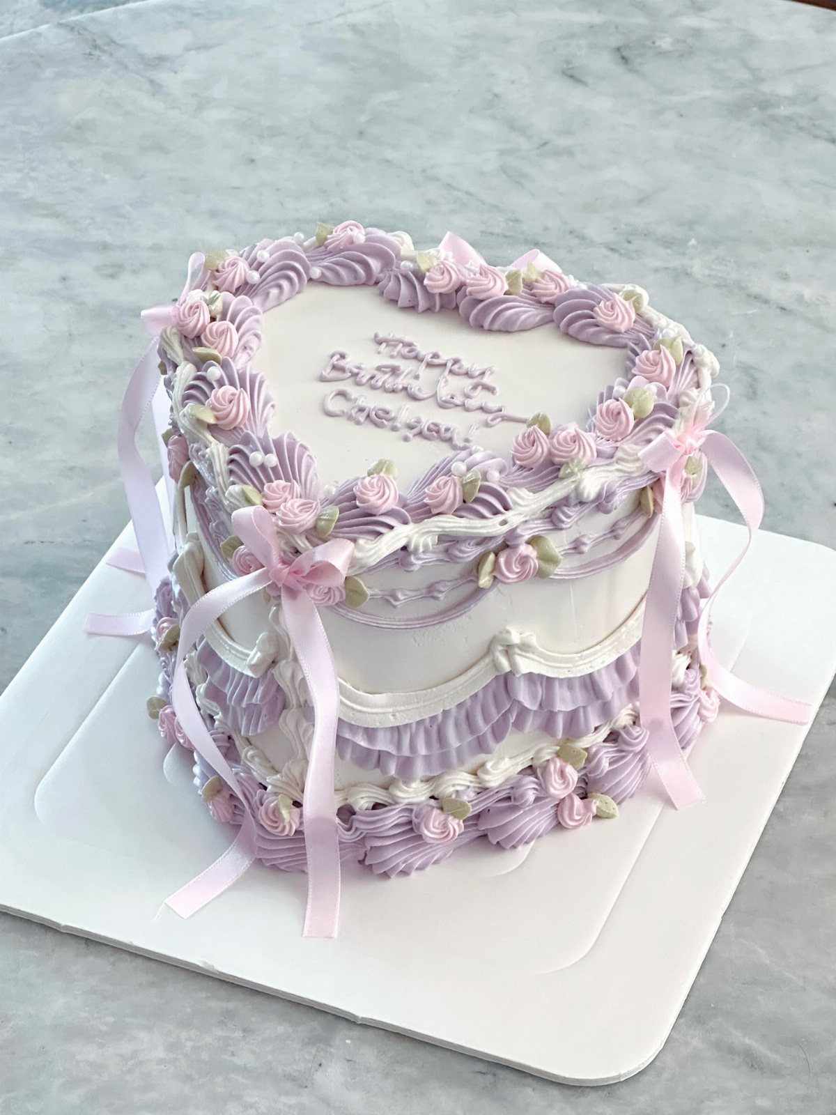 Heart-shaped white cake with purple frosting swirls, pink roses, and purple ribbons.