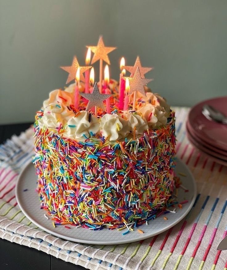 Sprinkle-covered birthday cake with lit pink candles and gold star toppers on a plate.