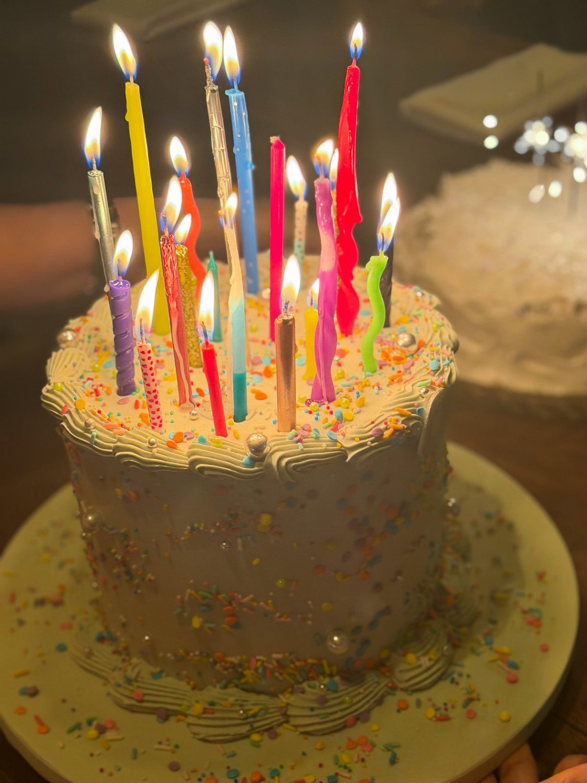 White frosted birthday cake with multicolored sprinkles and many lit rainbow candles.