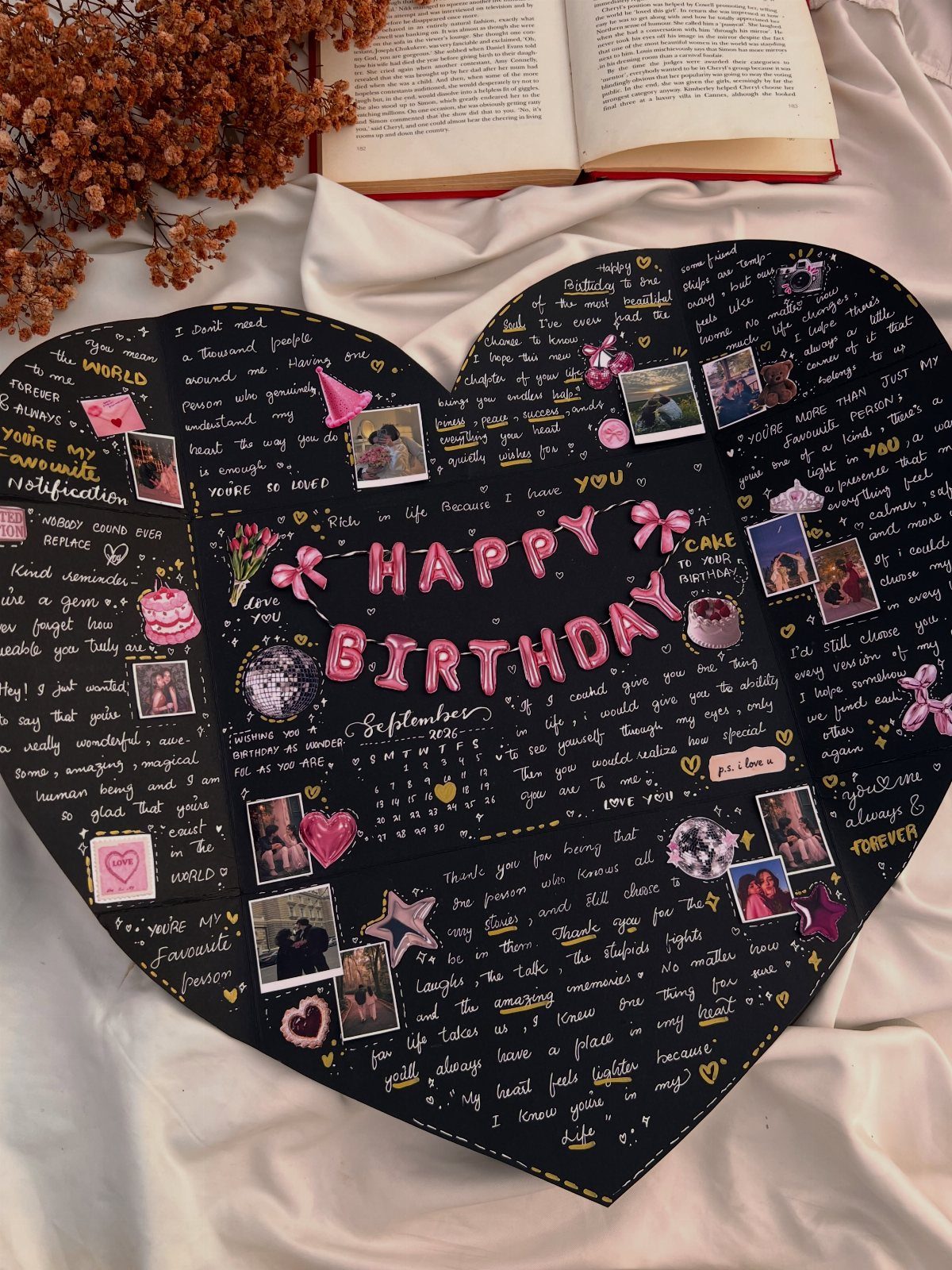Heart-shaped black birthday card with pink lettering, photos, and handwritten messages on white fabric.
