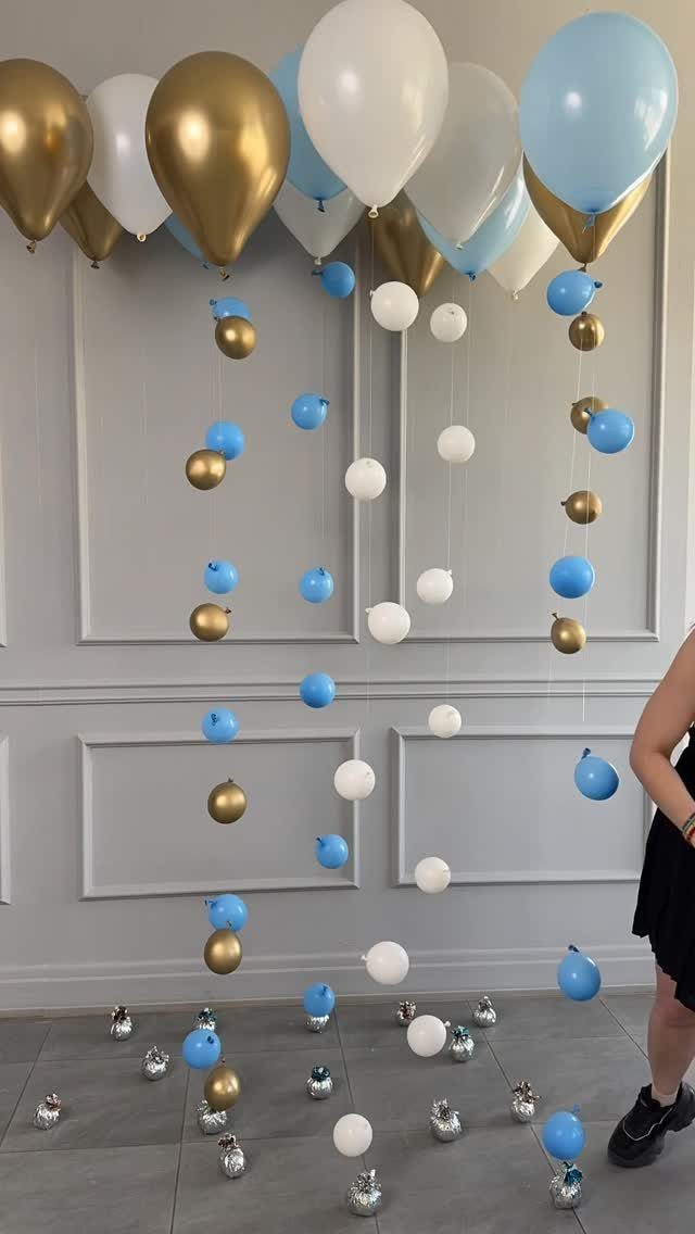 Gold, white, and blue balloons floating and hanging against a grey paneled wall.