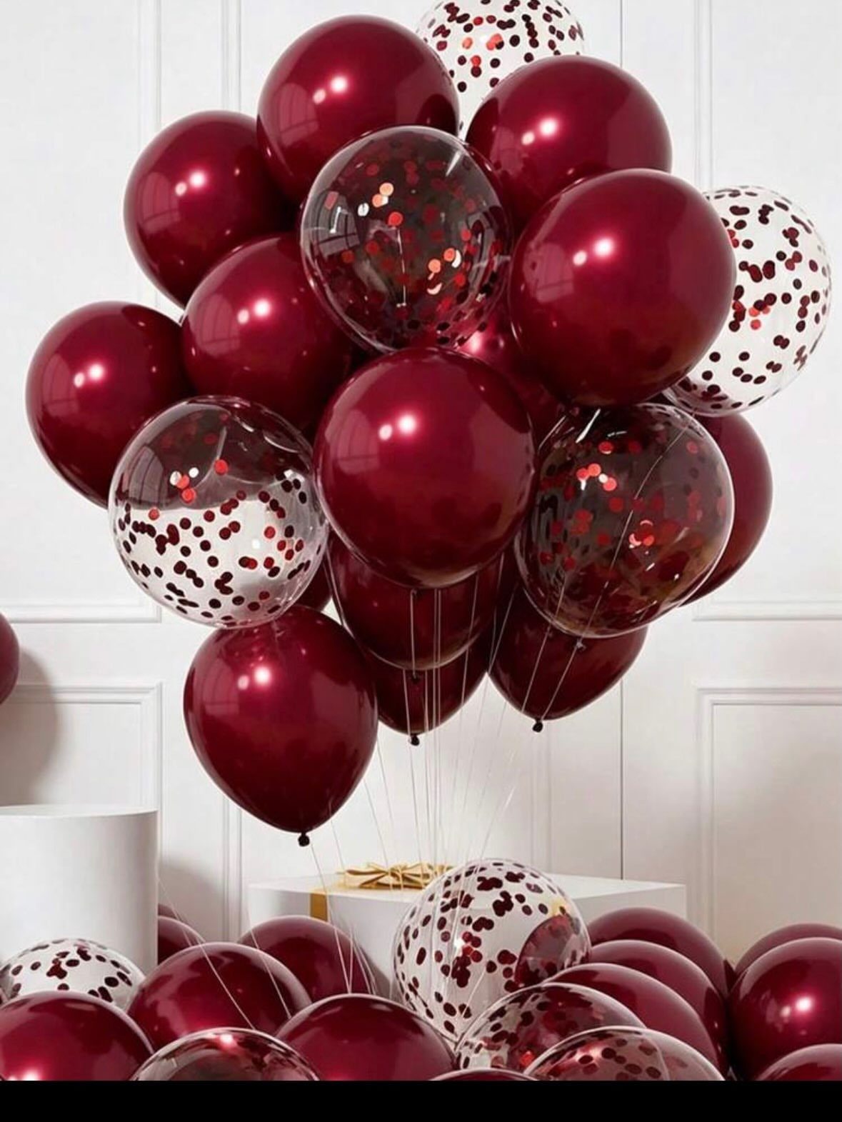 Burgundy metallic balloons and clear confetti-filled balloons floating against a white wall.