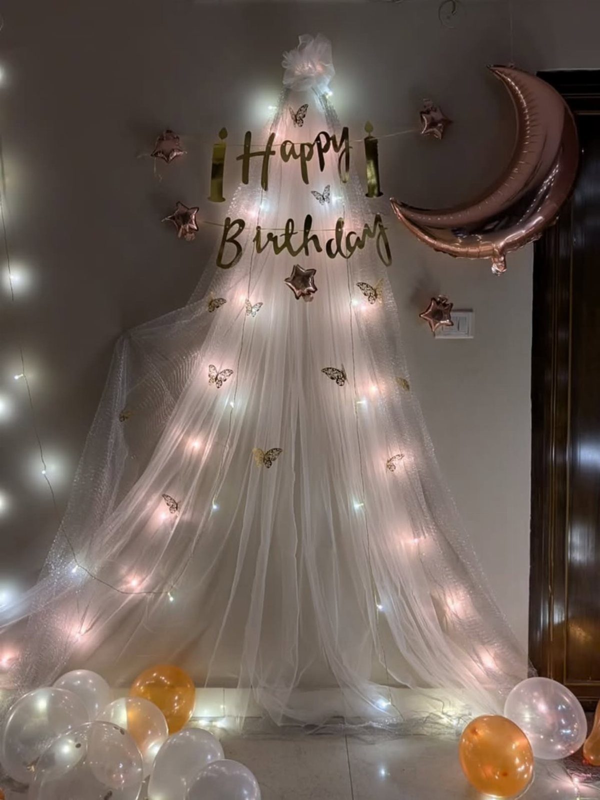 White sheer fabric backdrop with warm fairy lights, gold birthday lettering, and balloons.