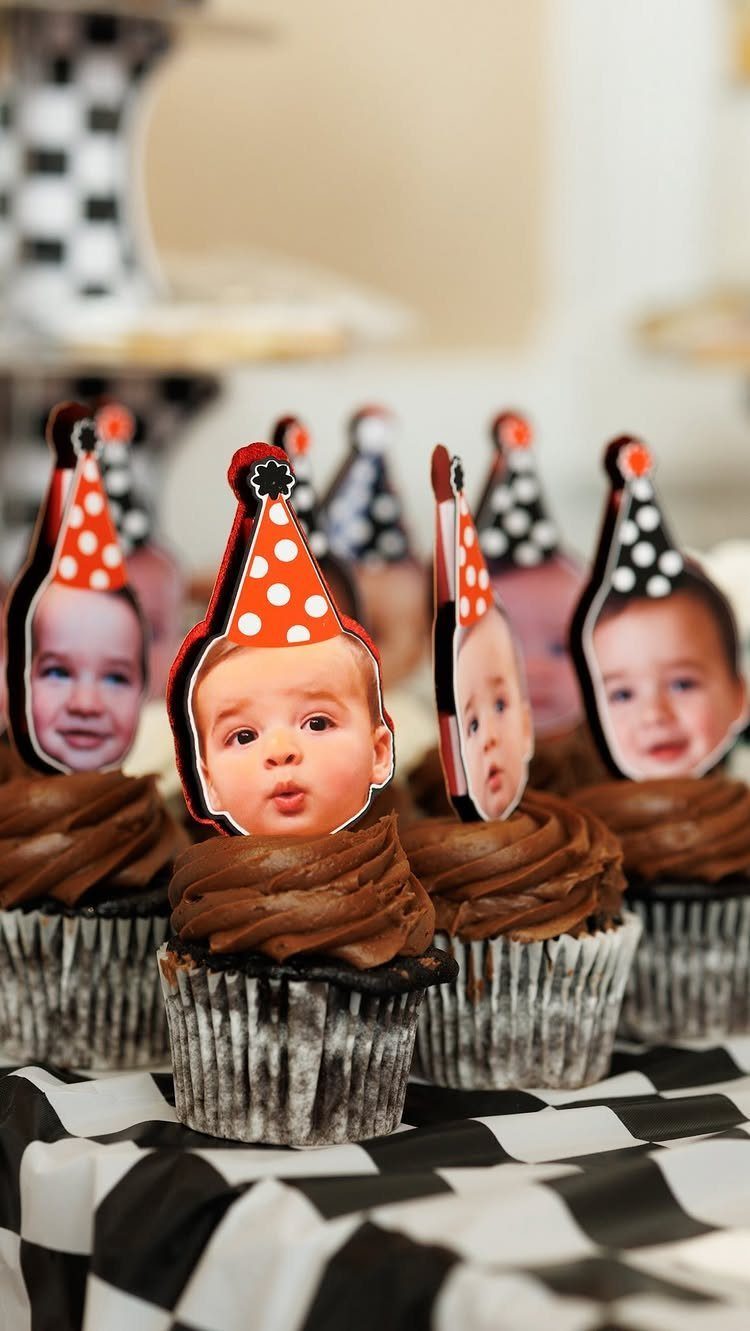 Chocolate cupcakes topped with custom baby face cutouts wearing party hats on checkered cloth
