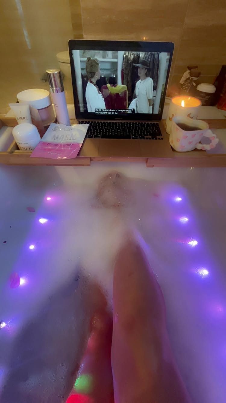 Wooden bathtub tray with laptop, candle, and skincare products over a bubbly bath.