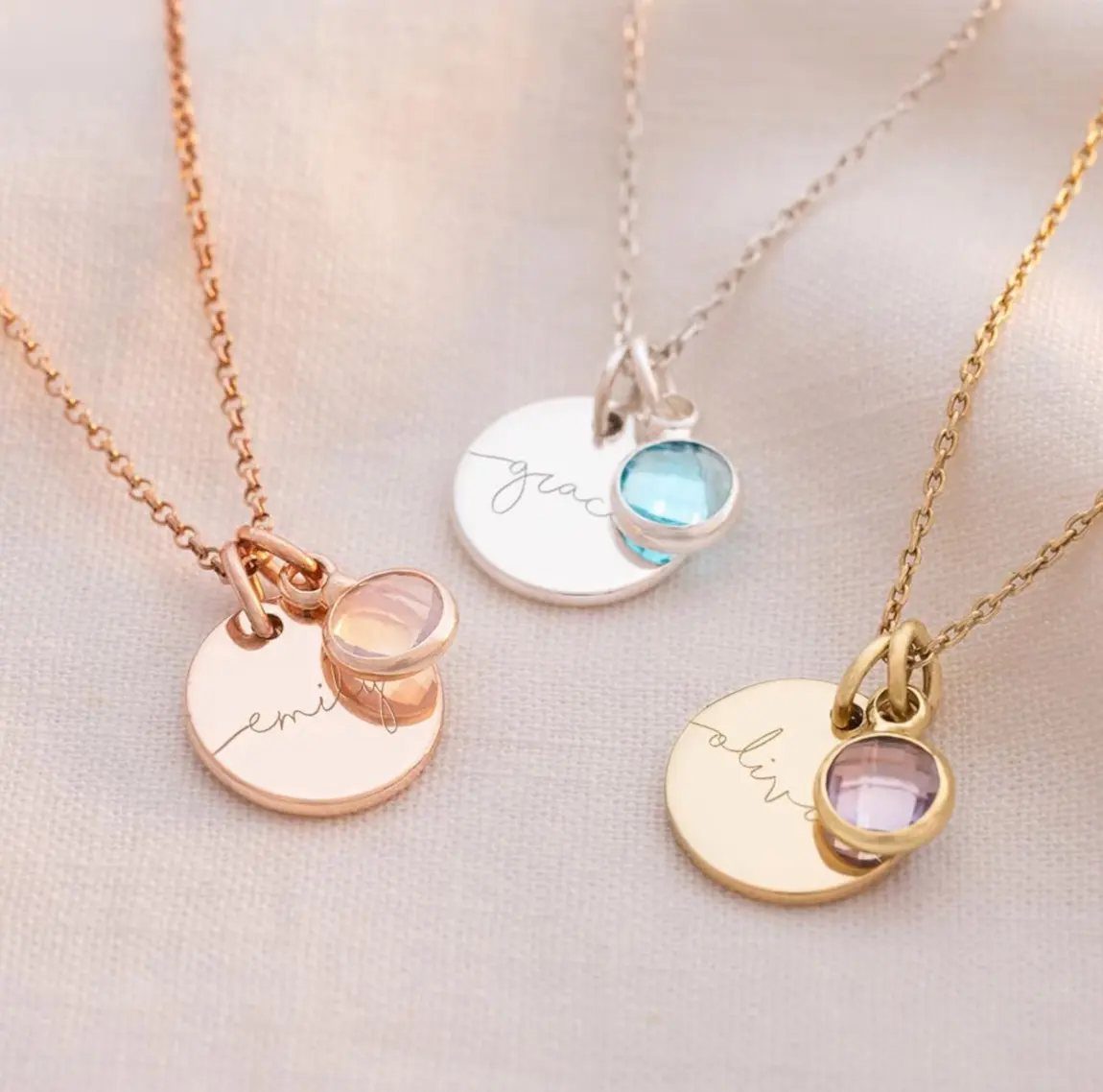 Three personalized necklaces with engraved name discs and colorful birthstone gems on white fabric