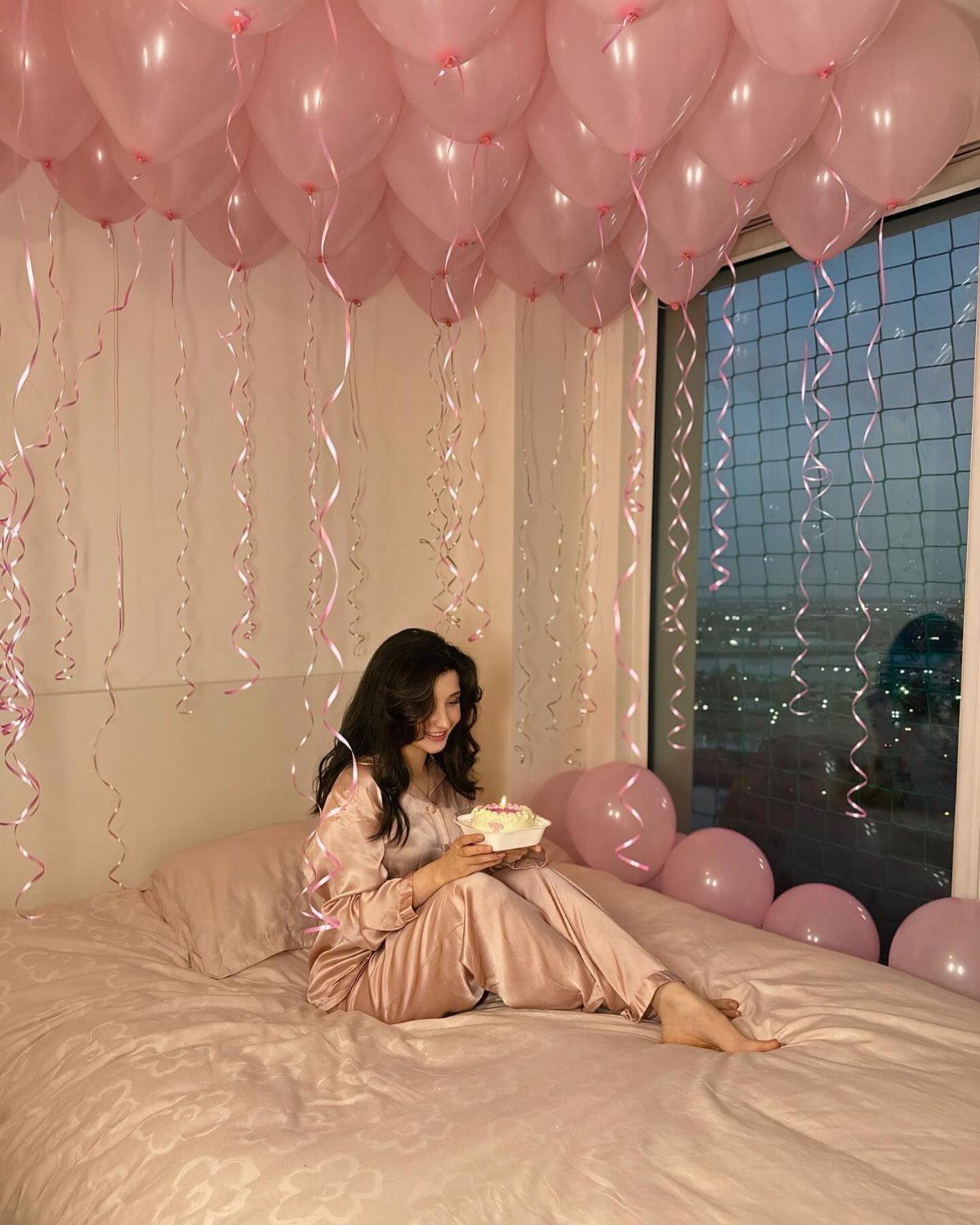 Woman in pink pajamas holding a birthday cake under pink ceiling balloons