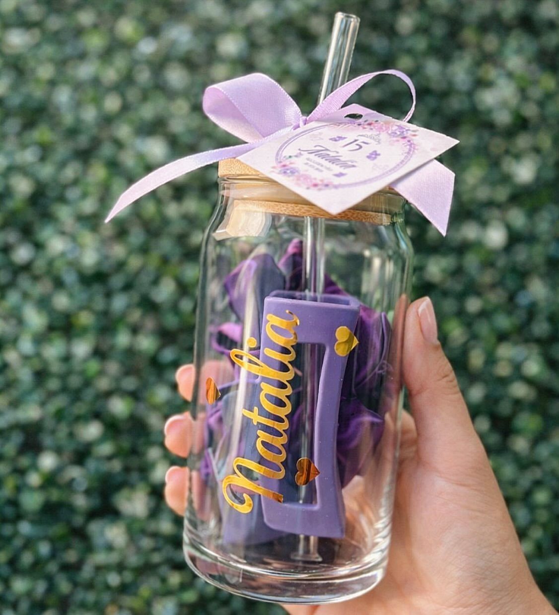 Glass tumbler jar with a purple ribbon and personalized name inside against greenery