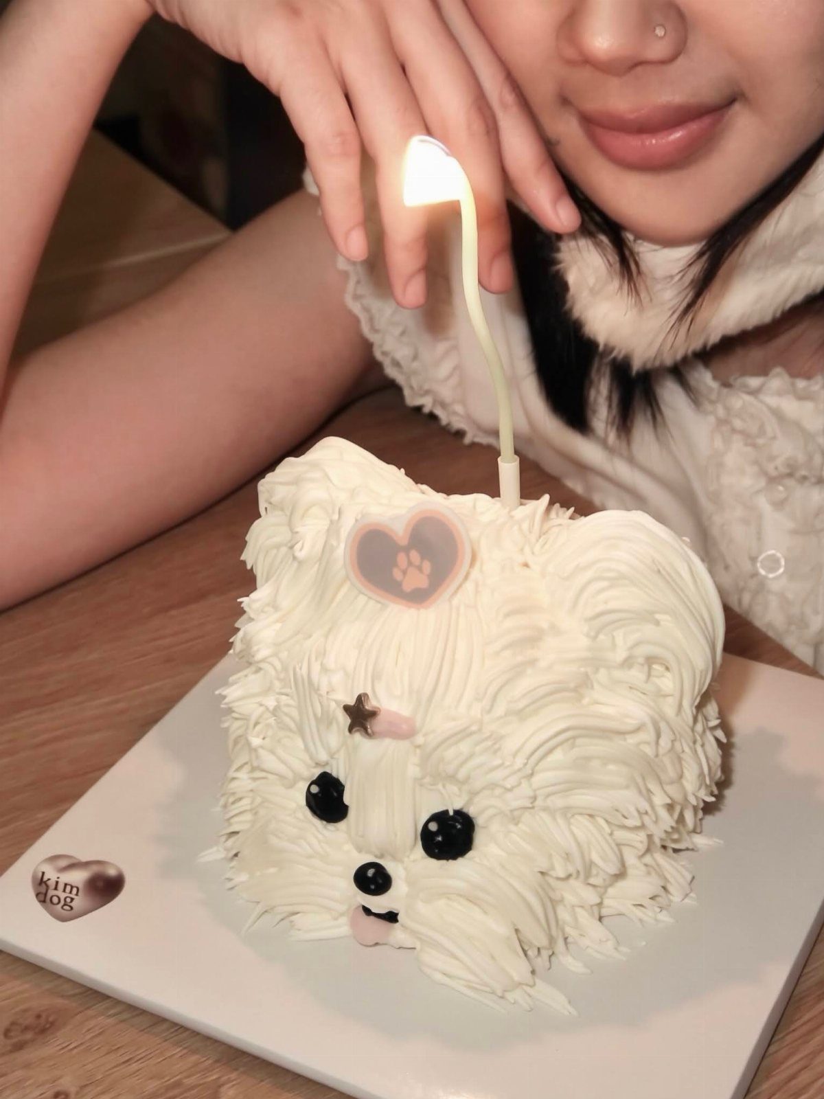 White dog-shaped birthday cake with a lit candle and a woman leaning over.