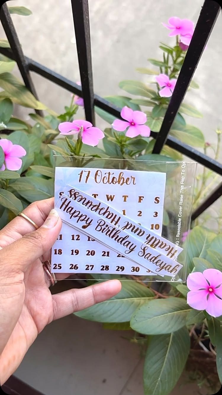 Acrylic birthday invitation card with gold text and October calendar held against pink flowers