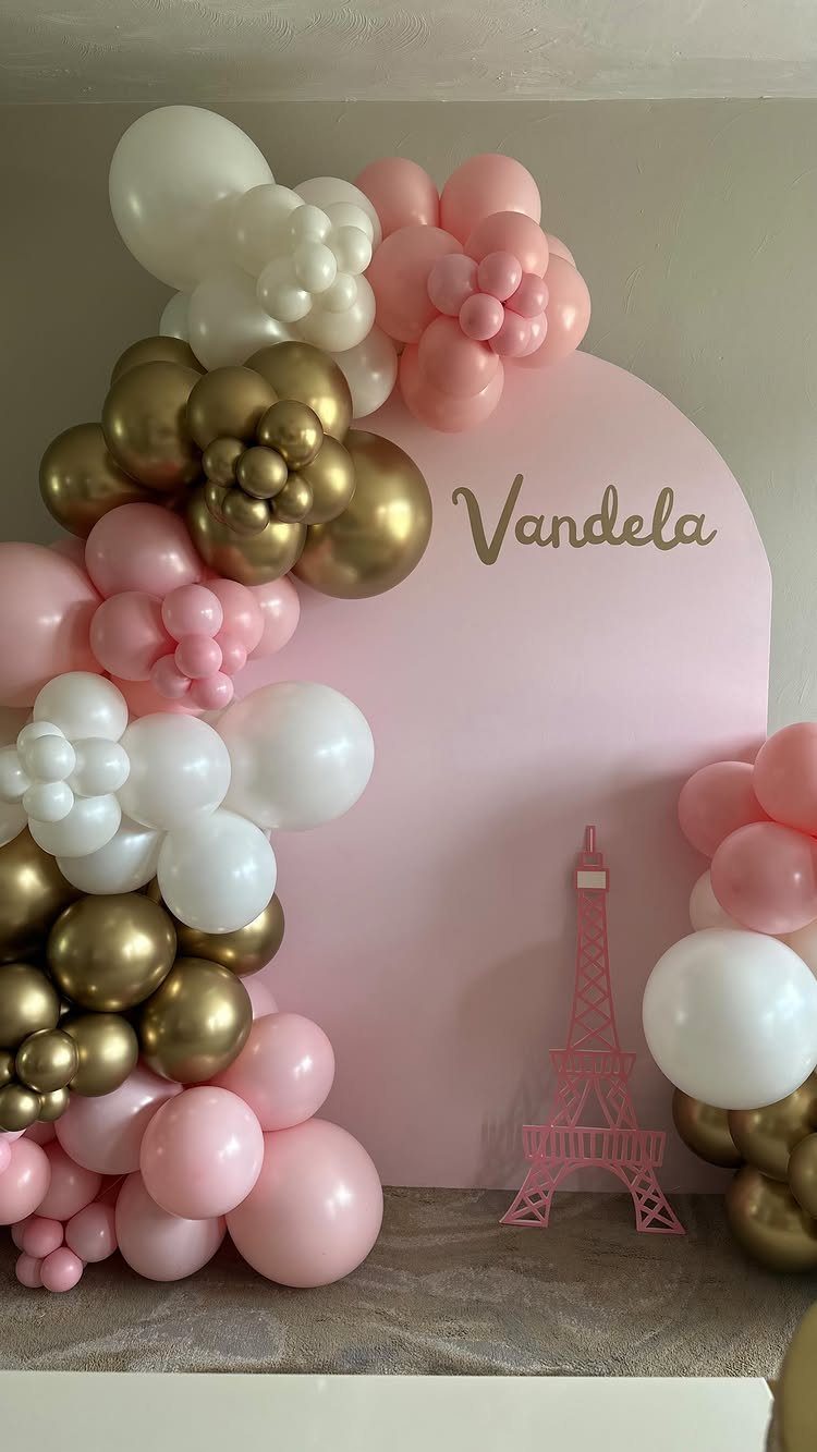 Pink arched backdrop with gold lettering, surrounded by white, pink, and gold balloons.